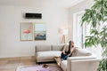 Black Bosch Climate 7000i inverter air conditioner mounted on a white wall in a modern living room with a gray sofa and large window sunlight