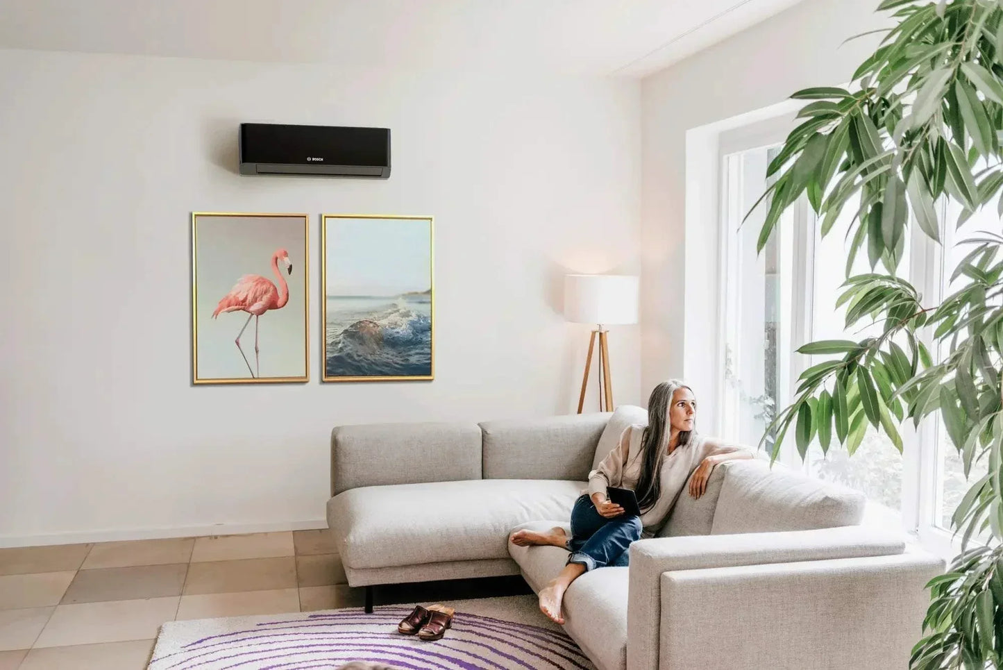 Black Bosch Climate 7000i inverter air conditioner mounted on a white wall in a modern living room with a gray sofa and large window sunlight